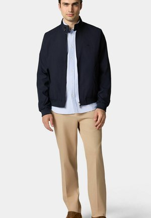 Navy bomberjack met een staand kraag, ritssluiting en geribde manchetten, gedragen over een lichtblauw button-up shirt, gecombineerd met beige broek.