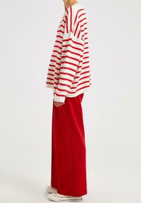 Person mit rot-weiß gestreiftem Langarmpullover, weiter roter Hose und weißen Turnschuhen vor einfarbigem Hintergrund.