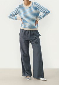 Pull rayé bleu et beige à manches longues, associé à un pantalon gris ample et des ballerines argentées, présentant un design minimaliste.