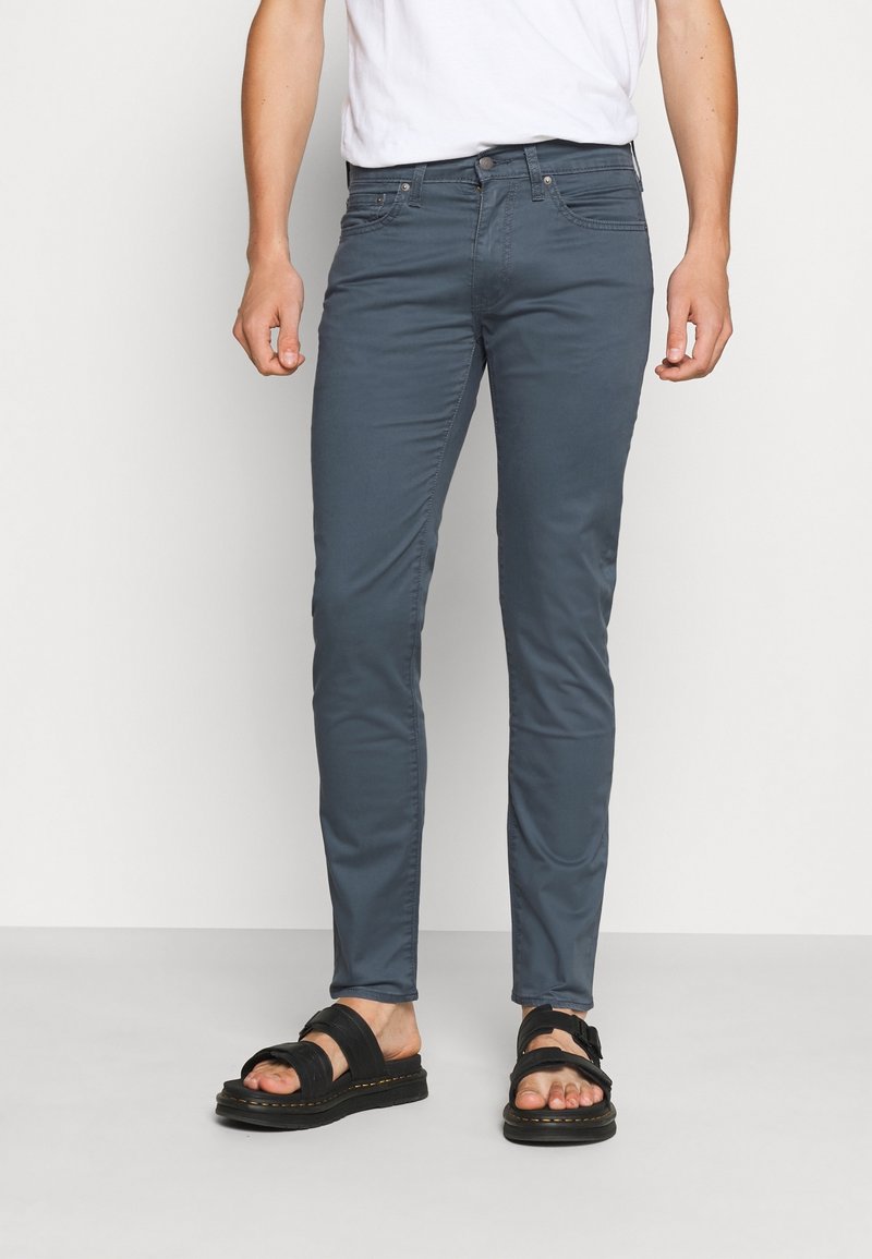 Homme portant un pantalon gris foncé ajusté, une chemise blanche et des sandales noires, se tenant sur un sol gris clair devant un fond blanc.