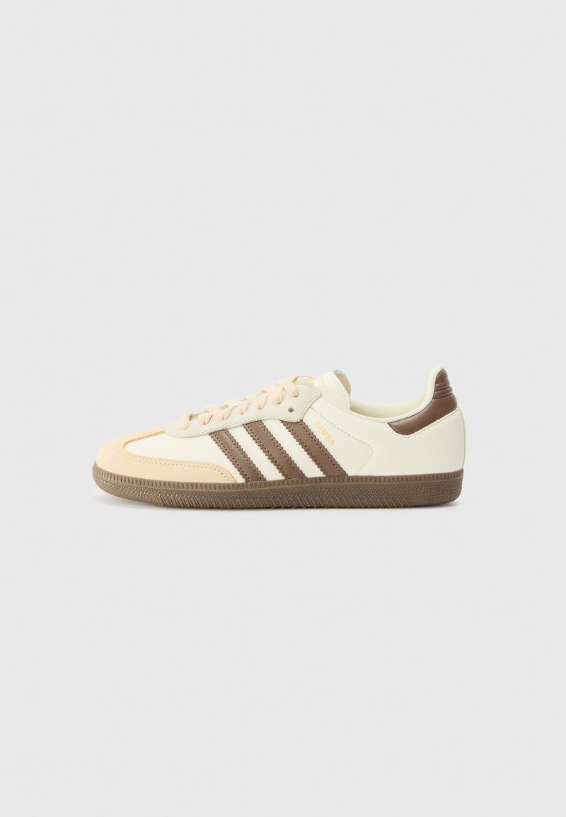 Des baskets avec un dessus beige, des accents marron clair, trois rayures marron foncé et une semelle en caoutchouc marron texturée. Marque clairement visible sur le côté.