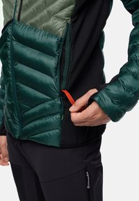 Grüne und schwarze isolierte Jacke mit einer puffigen Textur an der Vorderseite und glatten Ärmeln, mit einem kontrastierenden orangefarbenen Reißverschluss-Zipper.