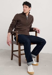 Rjava zip-up jopica, temno denim kavbojke, bele superge s svetlo rjavimi podplati. Model sedi na lesenem stolu pred nevtralnim ozadjem.