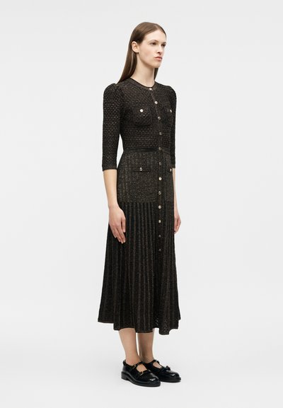 Γυναίκα που στέκεται φορώντας μαύρο υφασμάτινο μεσαίο φόρεμα με κουμπιά και μπροστινές τσέπες, συνδυασμένο με μαύρα παπούτσια Mary Jane, σε ανοιχτό γκρι φόντο.