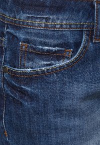 Jeans de mezclilla en un azul oscuro con una textura ligeramente desgastada. Presentan un bolsillo trasero con costuras naranjas y un detalle de botón de metal en el costado.