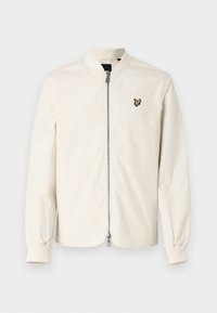 Lichtbeige bomberjack met een staande kraag, lange mouwen en een rits aan de voorkant. Heeft een geborduurd zwart en groen logo op de borst.