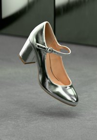 Silver metallic heel shoe with rounded toe, ankle strap, and block heel. Smooth texture with a glossy finish. Light brown sole.