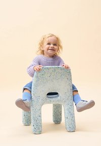 Kleurrijke, gespikkelde plastic stoel met afgeronde randen en uitsparing als handvat. Een kind zit erop, gekleed in een lichtpaarse trui en blauwe sokken.
