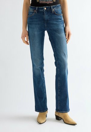 Persona che indossa jeans blu a gamba dritta e stivaletti beige in camoscio, in piedi davanti a uno sfondo bianco semplice.