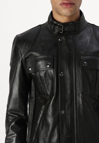 Blouson en cuir noir avec un col haut, deux poches poitrine avec boutons-pression, des accents aux épaules et une texture lisse.