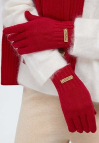 Rote gestrickte Handschuhe mit gerippten Bündchen und einem Etikett, getragen über einem weißen flauschigen Pullover und beigem Stoff. Die Textur ist weich und warm.