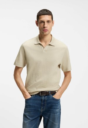 Young man with short hair wearing a textured beige short-sleeve polo and blue jeans with hands in pockets, plain white background.