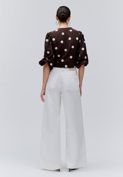 Naisel juuksed on kuklas, ta kannab pruuni täpilist pluusi pufvari varrukatega ning kõrge vöökohaga laiade valgete pükstega, seistes tavalise tausta ees.