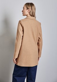 Abrigo de mezcla de lana de camello con mangas largas, cuello con muesca y corte recto, combinado con pantalones de mezclilla de pierna ancha de color azul oscuro.