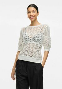 Bílý pletený svetr s vzorovaným designem, krátkými nabíranými rukávy a žebrovaným lemem, v kombinaci s černými skládacími kalhotami.