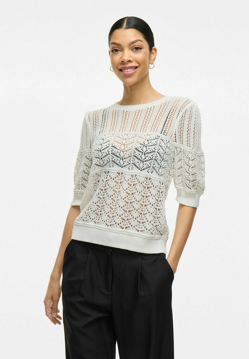 Bílý pletený svetr s vzorovaným designem, krátkými nabíranými rukávy a žebrovaným lemem, v kombinaci s černými skládacími kalhotami.