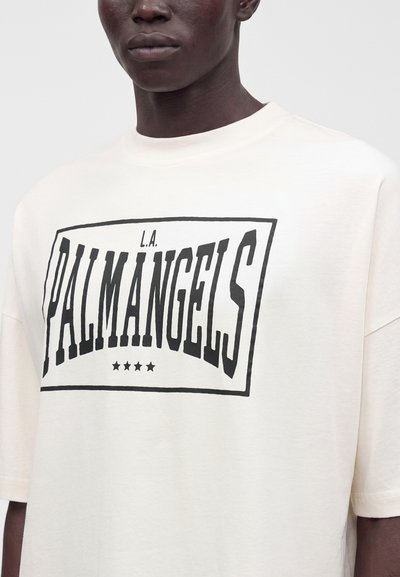 Άνδρας που φοράει λευκό T-shirt με μαύρα έντονα γράμματα "L.A. PALM ANGELS" και τέσσερα αστέρια μέσα σε ορθογώνιο πλαίσιο στο στήθος.