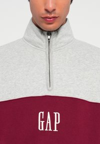 Gray and burgundy zip-up sweatshirt with a high collar; features white "GAP" logo on the lower part and a textured fabric.