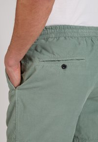 Mano de hombre en el bolsillo de pantalones de pana verde claro con cintura elástica y bolsillo trasero con botón, mostrado desde la cintura hasta la mitad del muslo.