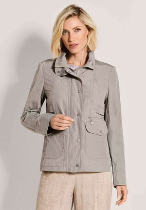 Blonde vrouw draagt een lichtgrijze jas met ritssluiting, knopen en zakken, gecombineerd met een beige broek, poseert tegen een effen achtergrond.