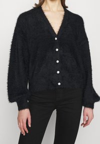 Cardigan noir et duveteux avec un col en V, des manches longues, et une fermeture à boutons comprenant six boutons perlés sur le devant. Texture douce.