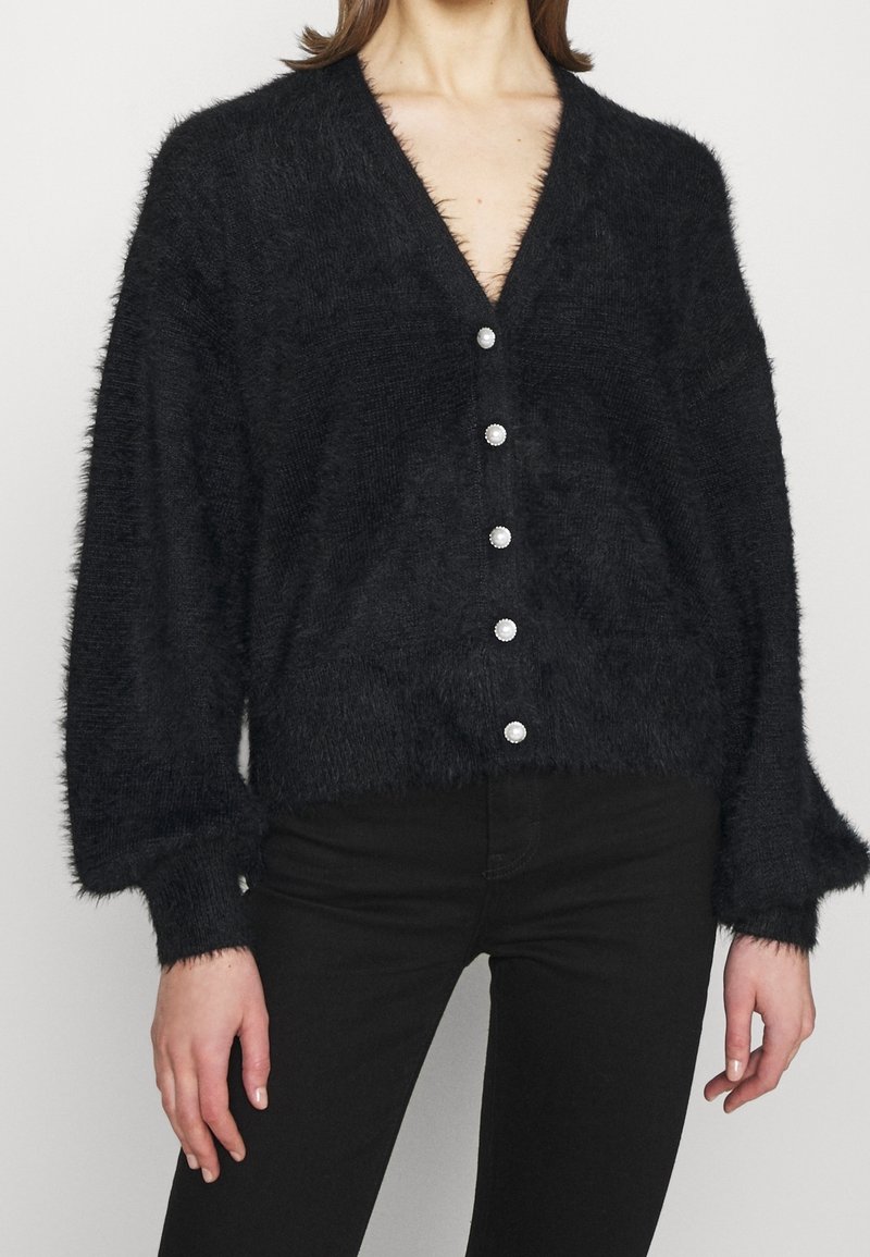 Cardigan noir et duveteux avec un col en V, des manches longues, et une fermeture à boutons comprenant six boutons perlés sur le devant. Texture douce.