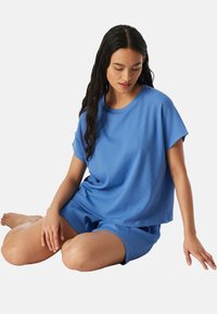 Junge Frau mit langen dunklen Haaren, sitzt im Schneidersitz und trägt ein lockeres blaues Kurzarmoberteil und passende Shorts vor weißem Hintergrund.