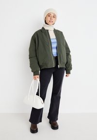 Jachetă bomber verde măslin peste un pulover crem cu gât înalt și un pulover albastru cu model. Jeans negri evazați, cizme maro cu platformă și o poșetă albă.