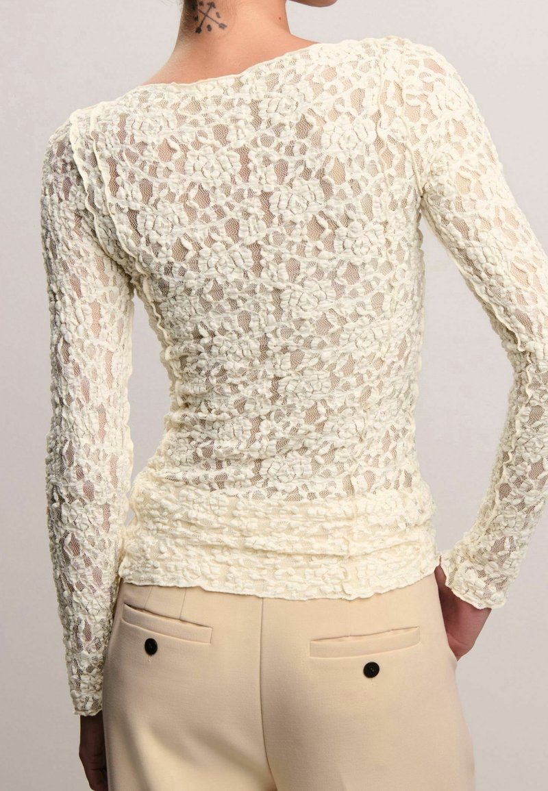 Femme portant un haut moulant en dentelle florale blanche à manches longues et un pantalon beige avec poches arrière et boutons noirs, vue de dos.