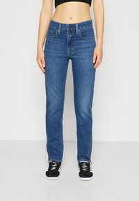 Jeans de mezclilla azul con corte recto, cierre de botón y bolsillos frontales. Combinados con zapatillas negras. Textura suave.