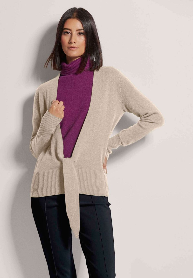 Beige gestrickte Strickjacke mit Wickeldesign und Bindegürtel in der Taille, kombiniert mit einem figurschmeichelnden lila Rollkragenpullover und schwarzen Hosen.