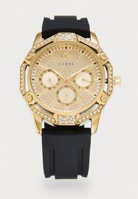 Montre Guess dorée avec un cadran et une lunette ornés de cristaux, bracelet en silicone noir, trois sous-cadrans pour les fonctions 24 heures, jour et date.