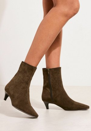 Brązowe zamszowe botki za kostkę z bocznymi zamkami i niskimi obcasami typu kitten, noszone na skrzyżowanych nogach na jednolitym tle.