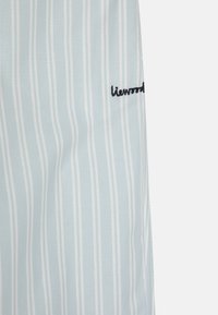 Lichtblauwe stof met verticale witte strepen. Kleine geborduurde "liewood" label in donkerblauwe draad aan de rechterzijde. Glad textuur.