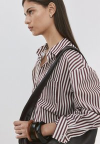 Mujer con cabello largo y oscuro lleva una camisa de rayas verticales en burdeos y blanco, lleva un bolso de cuero negro colgado del hombro, y usa pendientes de aro y pulseras.