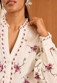 Blusa bianca con ricami floreali in rosa e verde. Presenta un colletto arricciato e chiusura con bottoni. Maniche lunghe a sbuffo con tessuto arricciato.