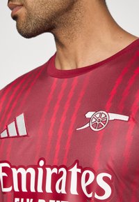 Rood atletisch shirt met korte mouwen, met diagonale strepen, een ronde hals en logo's van Emirates en Arsenal in contrasterend wit.