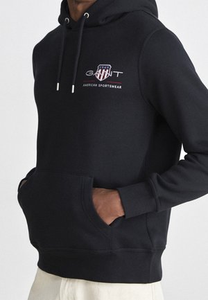 Czarna bluza z kapturem, z przednią kieszenią i sznurkami, z czerwono-białym logo w kształcie tarczy z napisem "GANT 1949" oraz "AMERICAN SPORTSWEAR" na piersi.