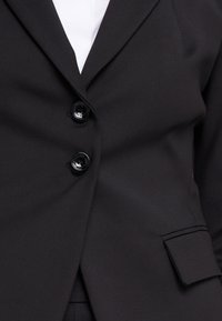 Svart kavaj med en slät textur, två knappar och en enkel ficka med klaff. Skräddarsydd design med slagkrage och en strukturerad silhuett.