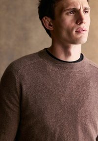 Brauner Wollpullover mit einem gerippten Halsausschnitt und Schulternähten. Weiche Textur und figurbetontes Design. Der Hintergrund hat einen neutralen Ton.