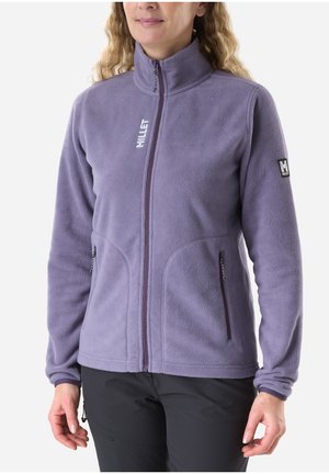 Woman wearing a lavender zip-up fleece jacket with two zippered side pockets and black pants, standing against a plain background.