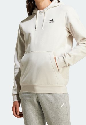 Personne portant un sweat à capuche crème Adidas avec poche avant, assorti à un pantalon de jogging Adidas gris clair, main dans la poche du sweat.