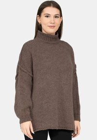 Vrouw draagt een loszittende bruine gebreide coltrui met lange mouwen en een zwarte broek, staand tegen een effen achtergrond.