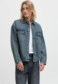 Denim jakna u tamnoplavoj boji s preklopom na dugmad, dva džepa na grudima i opuštenim oblikom. Nosiva preko bijelog majice i u kombinaciji s tamnim trapericama.