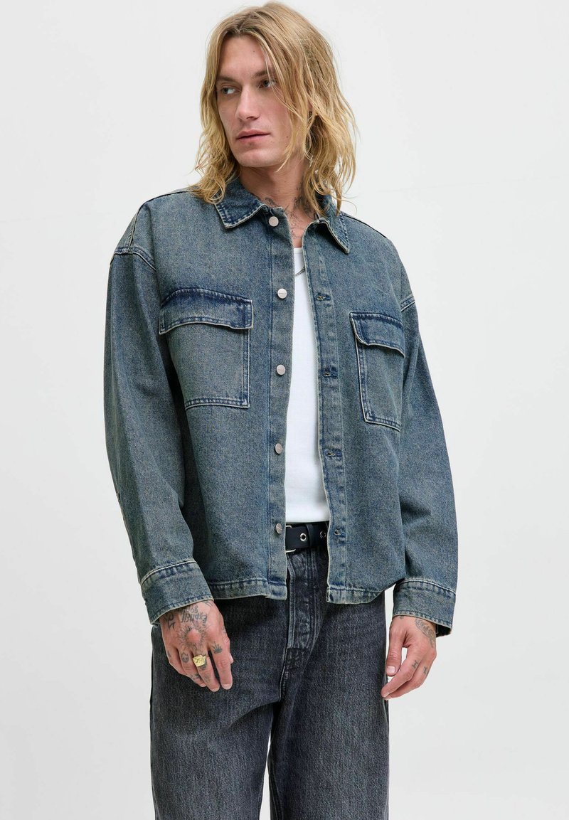 Denim jakna u tamnoplavoj boji s preklopom na dugmad, dva džepa na grudima i opuštenim oblikom. Nosiva preko bijelog majice i u kombinaciji s tamnim trapericama.