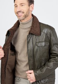 Grüner Lederjacke mit braunem Shearling-Kragen, Reißverschluss vorne, Seitentaschen und strukturierter Oberfläche, getragen über einem beigen Rollkragenpullover.