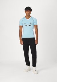 Ανοιχτό γαλάζιο αθλητικό μπλουζάκι με μαύρες λεπτομέρειες, έχει V-λαιμόκοψη, κοντά μανίκια και την επιγραφή "URTRN." Συνδυασμένο με μαύρες jogger παντελόνες και λευκά αθλητικά παπούτσια.