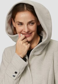 Veste zippée gris clair avec une grande capuche, tissu texturé et boutons-pression sur les poignets, présentant des accents argentés et une coupe décontractée.