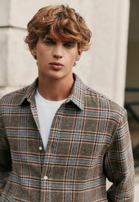 Bruin geruiten shirt met blauwe en rode accenten, knoopsluiting, zachte textuur en kraag. Draag dit over een wit shirt.