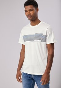 Bílý tričko s krátkým rukávem s vodorovným designem námořnické modré a světle modré pruhy na hrudi a textem pod ním. Měkký bavlněný materiál.